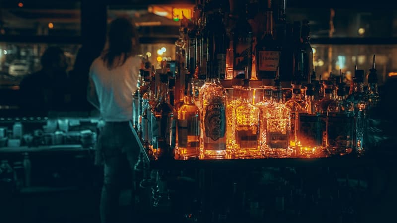 Scaffali di bottiglie di liquori retroilluminate brillano dietro un barista in un locale buio