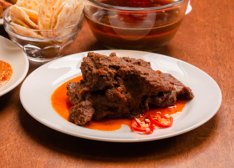 Un plato de rendang de ternera oscuro con guindilla roja en rodajas, roti canai y sambal detrás