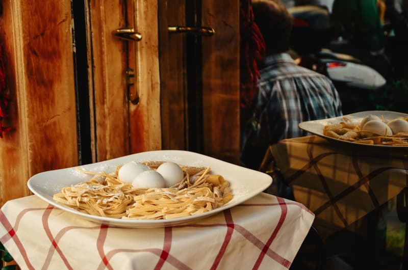 Tagliatelle fresche, rigatoni e uova su un piatto posato su una tovaglia a quadri davanti a una trattoria romana