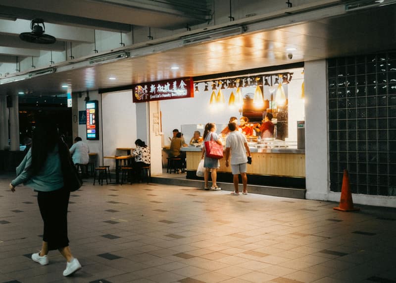 Hell erleuchteter Essensstand in Singapur bei Nacht, mit bestellenden Kunden an der Theke und Gästen auf Hockern