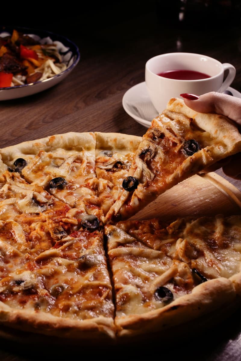 Una mano levantando una porción de pizza de queso y aceitunas en una mesa de restaurante informal