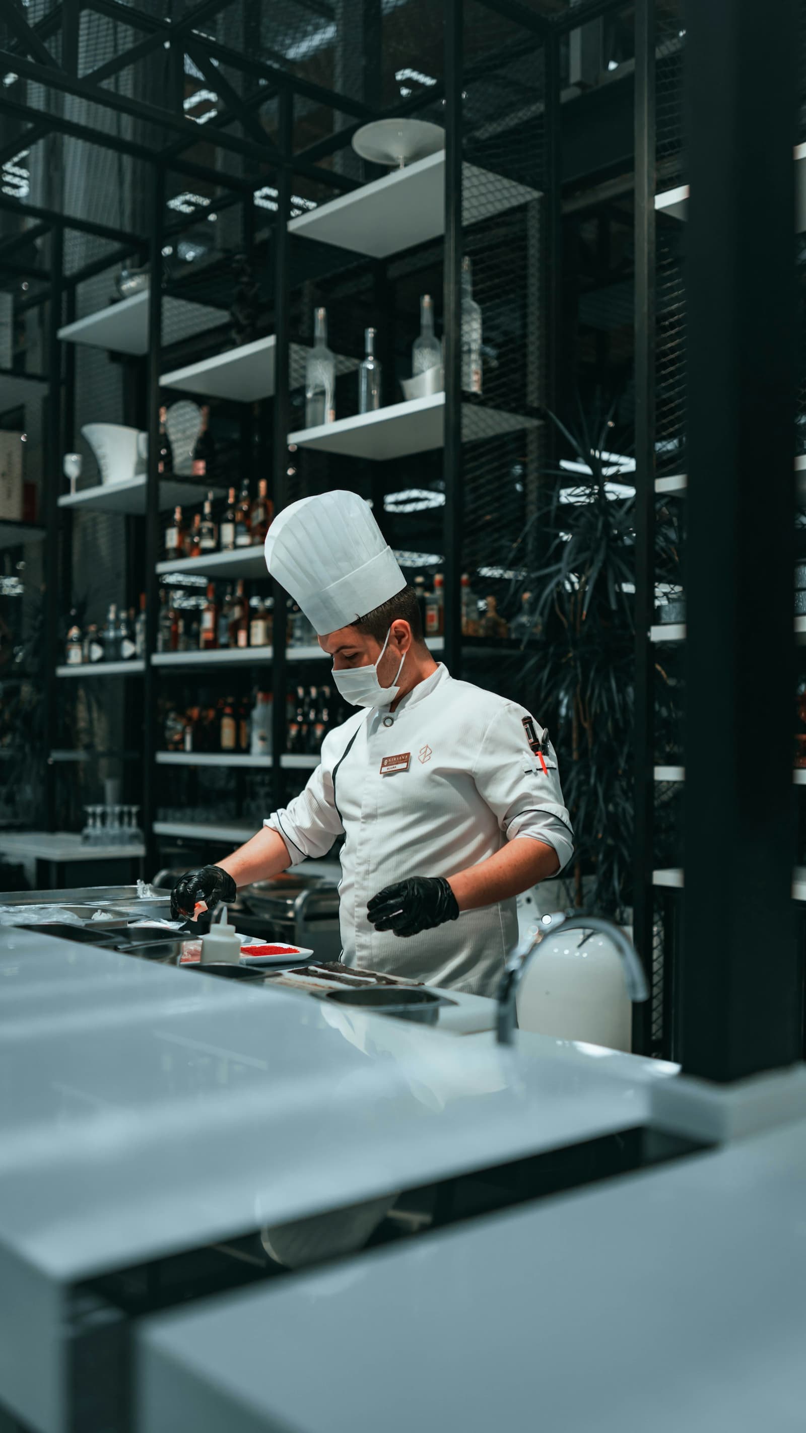 Chef in tall white toque plating a dish in a dimly lit restaurant kitchen