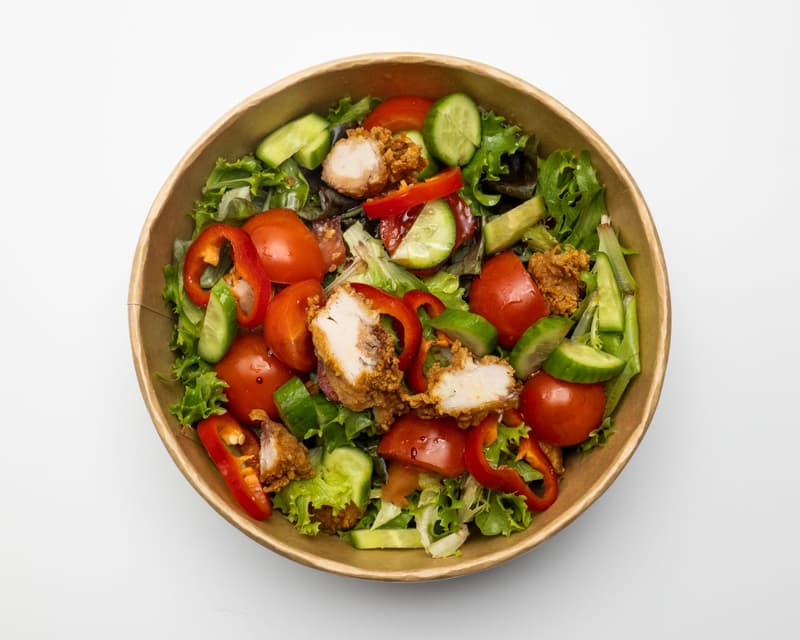 A wooden bowl of chicken salad with lettuce, tomatoes, peppers and cucumber