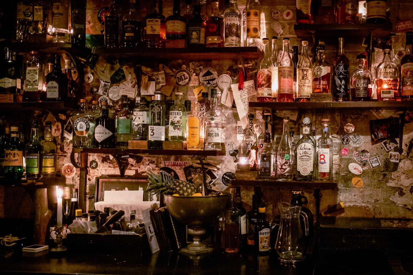 Cluttered back bar shelf of spirits bottles and stickers in a dimly lit speakeasy-style bar