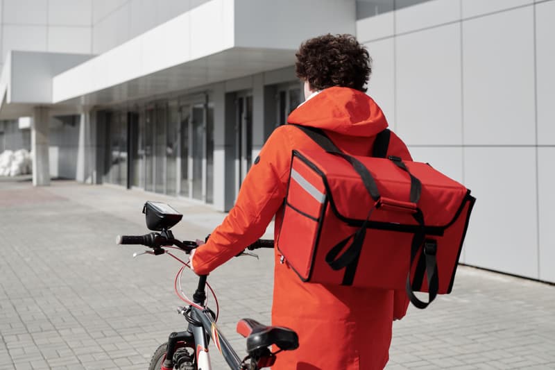 Rider con giacca rossa che cammina accanto alla bicicletta con una borsa termica in strada