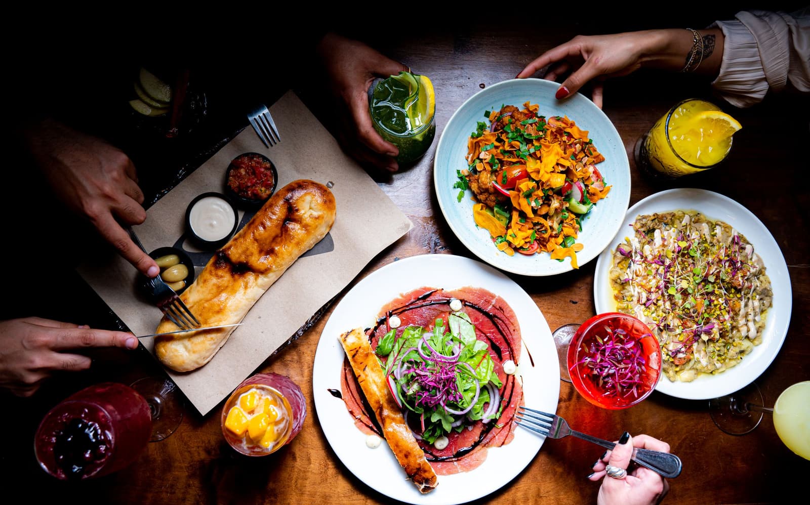 Shared Mediterranean lunch of salads, carpaccio, flatbread and colourful drinks on a wooden table