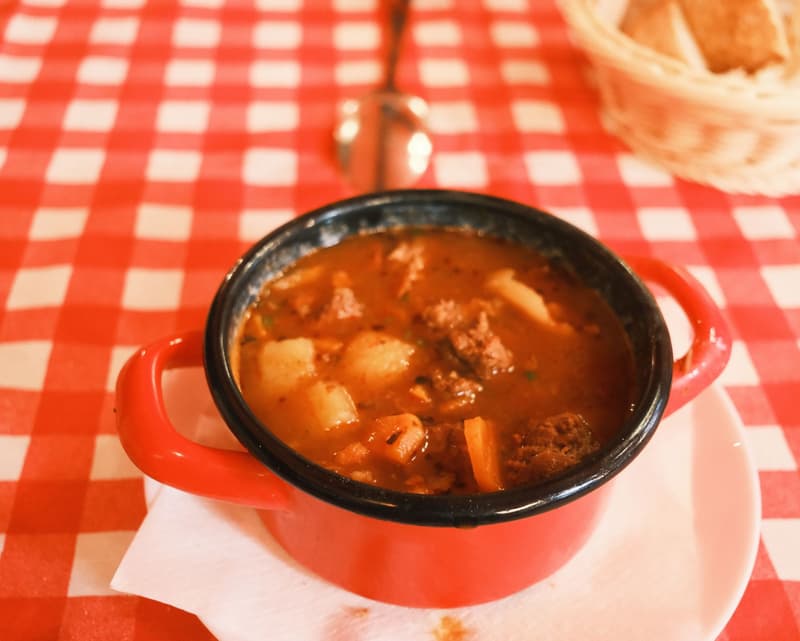 Ungarische Gulaschsuppe mit Rindfleisch, Kartoffeln und Karotten in einem roten Emailtopf auf karierter Tischdecke