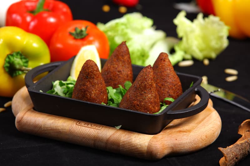 Conos de kibbeh frito servidos en una bandeja con hierbas frescas y pimientos de colores