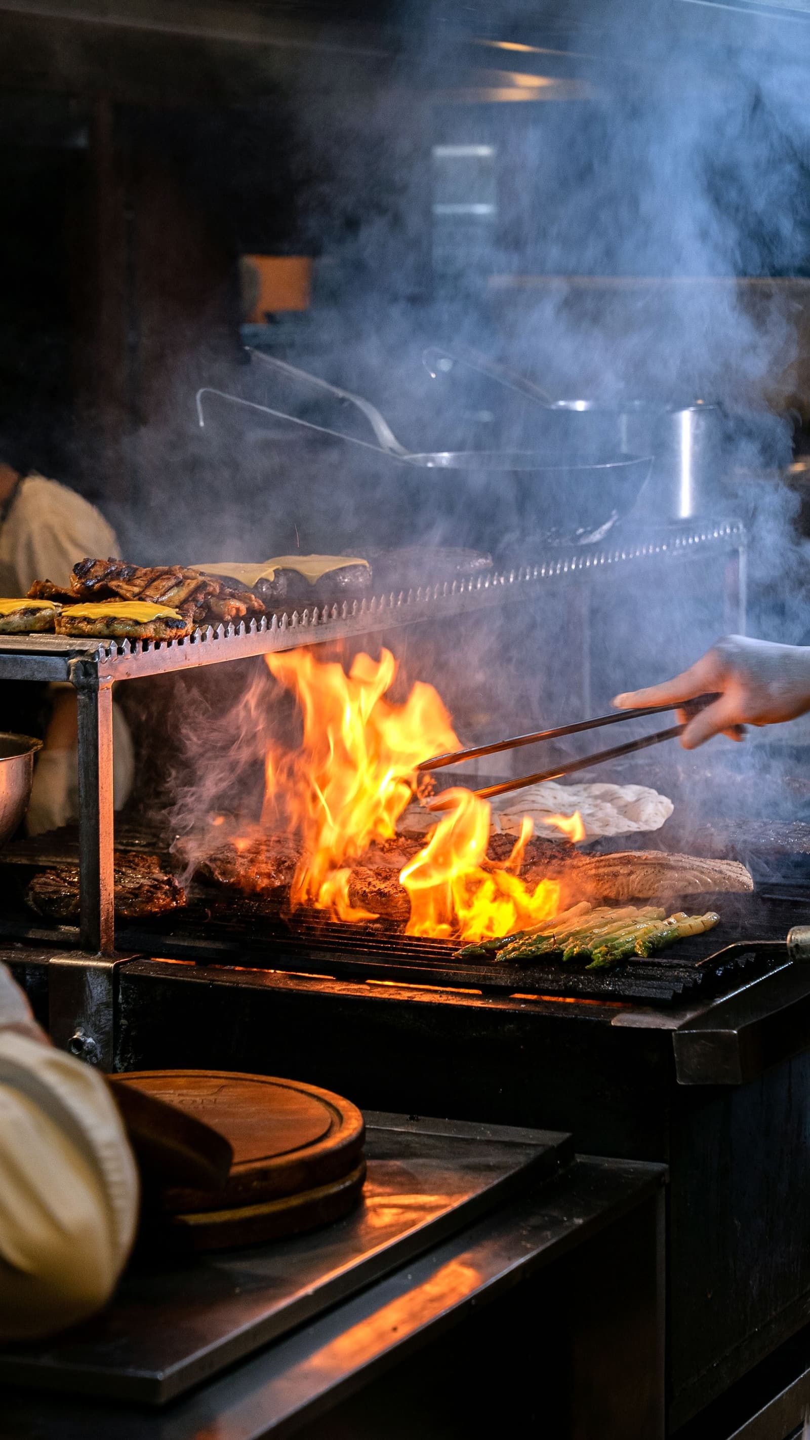 Chef grilling meat and asparagus over an open flame on a charcoal grill