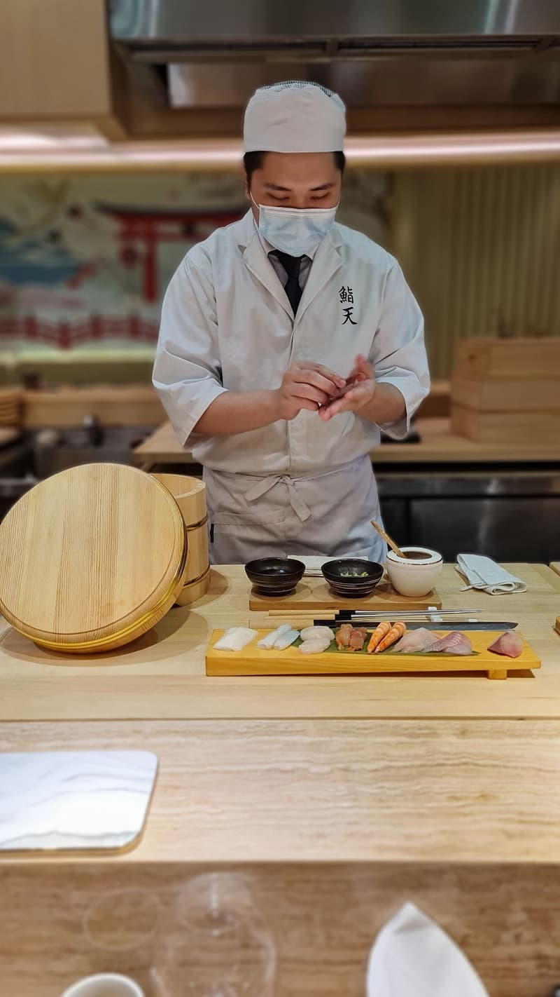 Ein Sushi-Koch mit Maske bereitet ein Tablett Nigiri und Sashimi an einer Holztheke zu
