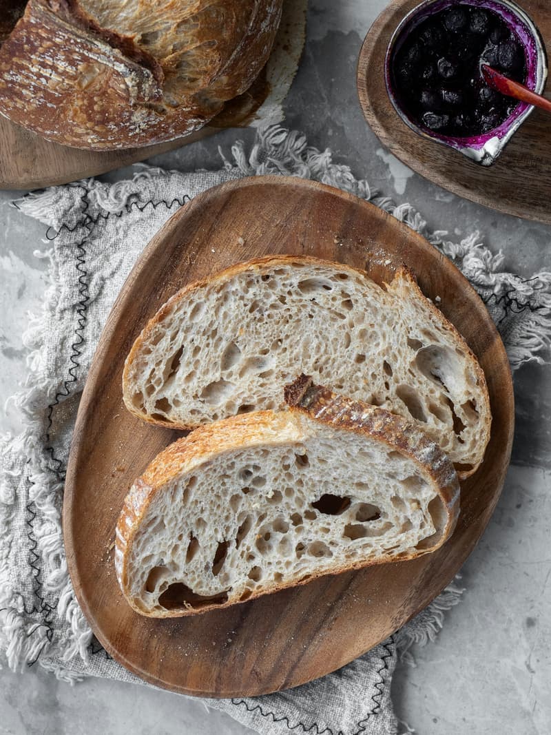Ein rundes, bemehltes Brot aufgeschnitten neben zwei Scheiben auf einem Holzbrett
