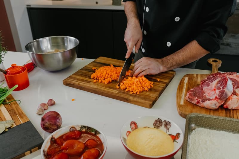 Un chef coupe des carottes sur une planche en bois près de viande crue et de tomates