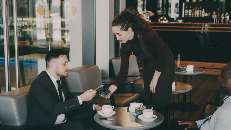 Cameriera che porge un POS a un cliente seduto al tavolo di un bar