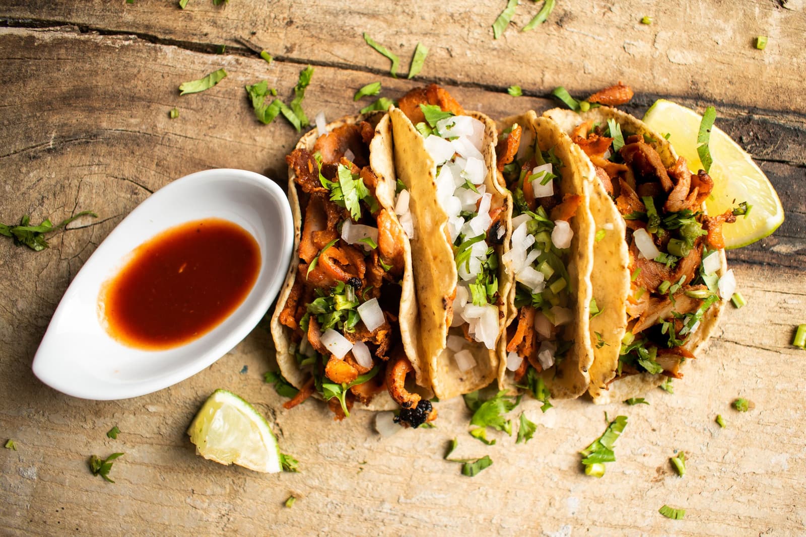 Quatre tacos al pastor avec oignon, coriandre, citron vert et salsa rouge sur une planche en bois