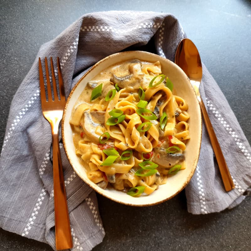 Ciotola di tagliatelle con salsa cremosa ai funghi e cipollotto, con forchetta e cucchiaio accanto