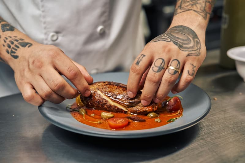 Manos tatuadas de un cocinero colocando una pieza de carne en un plato gris con salsa