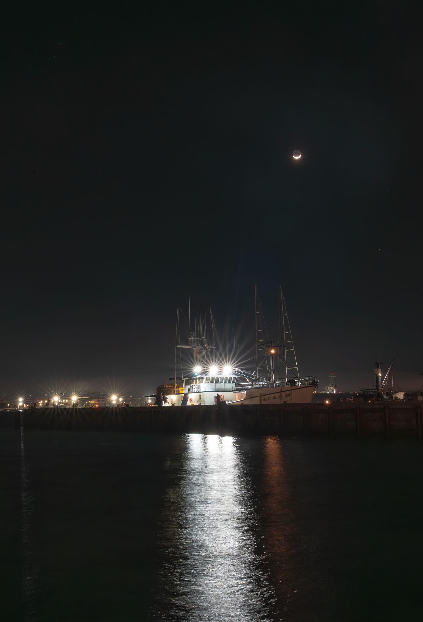 Beleuchtete Fischerboote in einem Thunfischhafen bei Nacht unter zunehmendem Mond