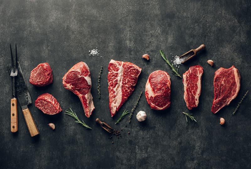 Steaks de bœuf crus de différentes pièces sur un fond sombre avec herbes et ail
