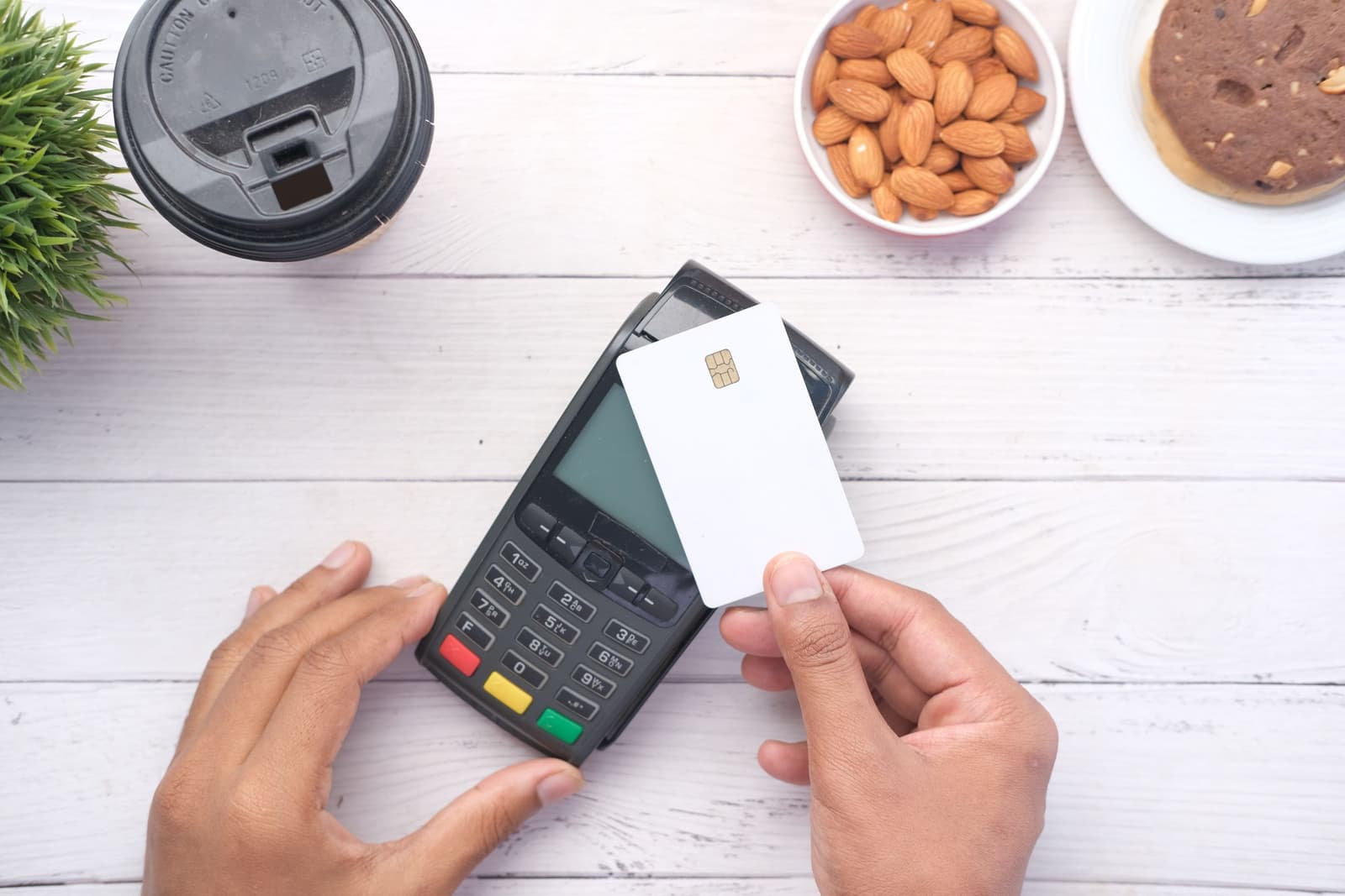 Des mains approchent une carte bancaire d'un terminal de paiement sur une table blanche, près d'un café à emporter et d'un bol d'amandes