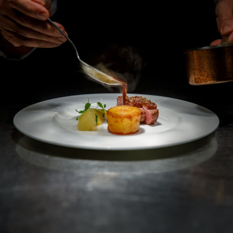 Ein Koch gießt Sauce über ein Fleischgericht in einer dunklen Fine-Dining-Küche
