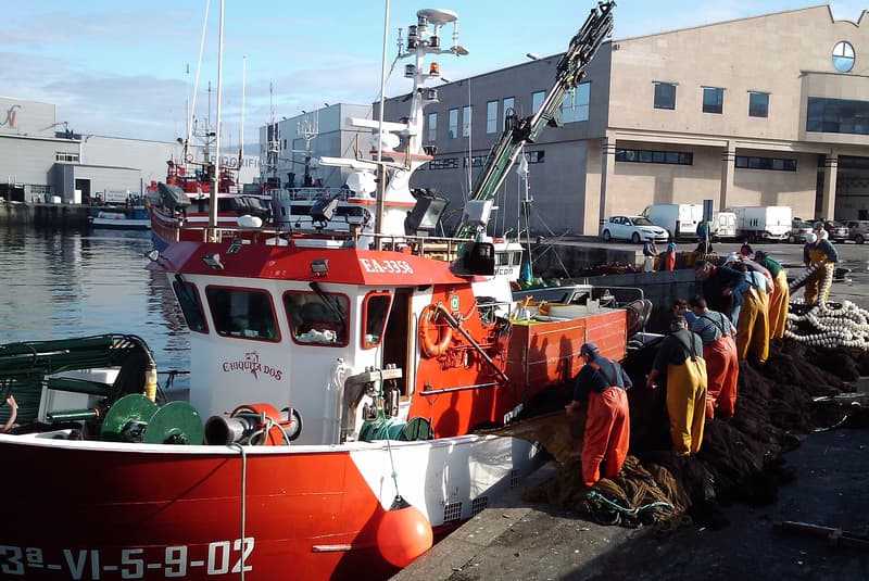 Pesquero rojo amarrado en el puerto de Vigo mientras marineros repasan redes en el muelle