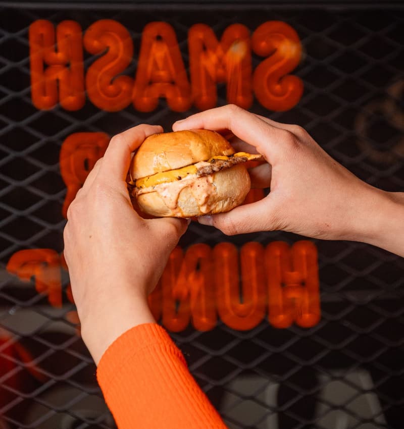 Des mains tiennent un smash burger au fromage fondu devant une enseigne au néon