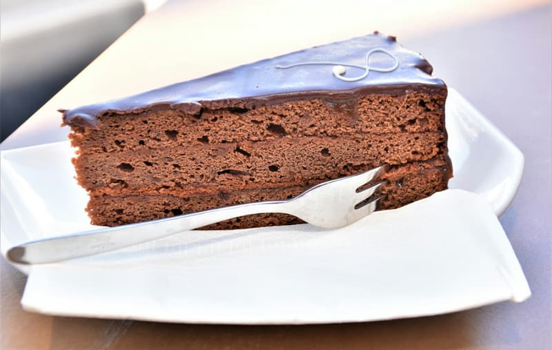 Stück Sachertorte mit glänzender Schokoladenglasur und einer Gabel auf einem weißen Teller