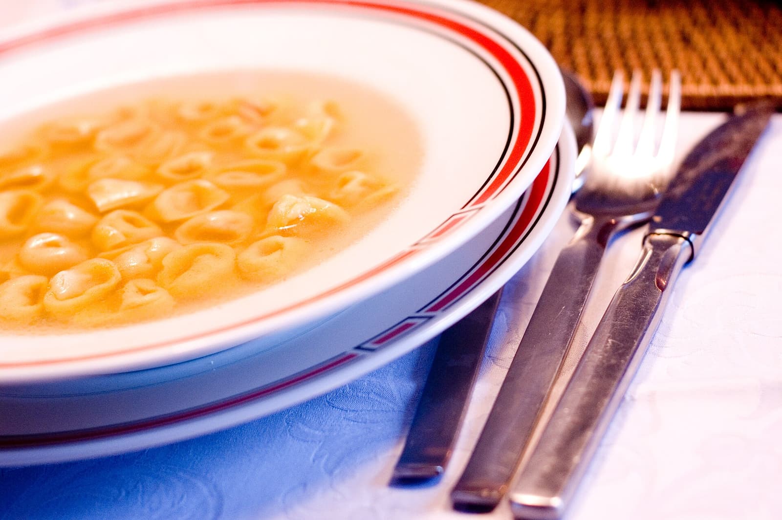 Ein weißer Teller mit kleinen ringförmigen Tortellini in goldener Brühe neben Gabel und Messer